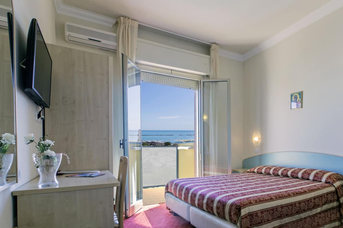 A hotel room with a bed, a desk, a TV, a window with a view of the beach, and a small religious icon on the wall.