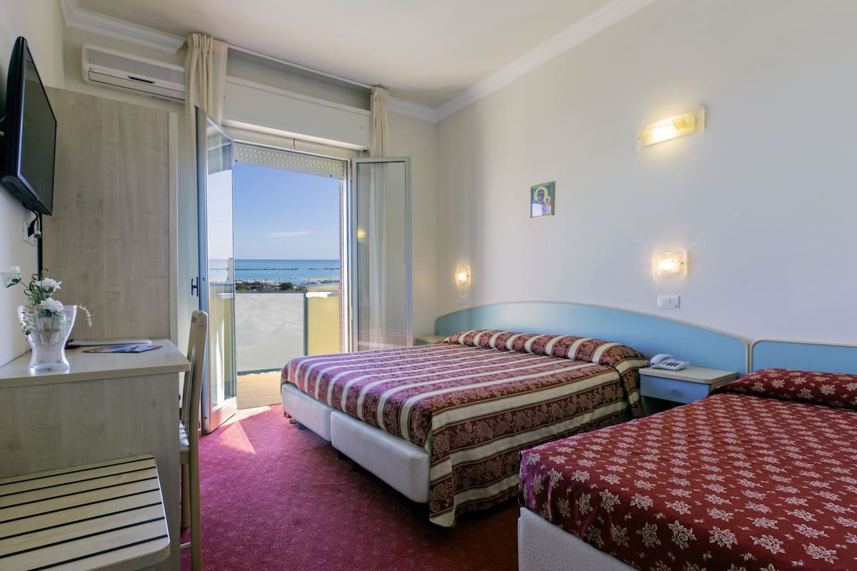 A hotel room with two beds, a TV, a desk with a vase of flowers, and a balcony with a view of the sea.