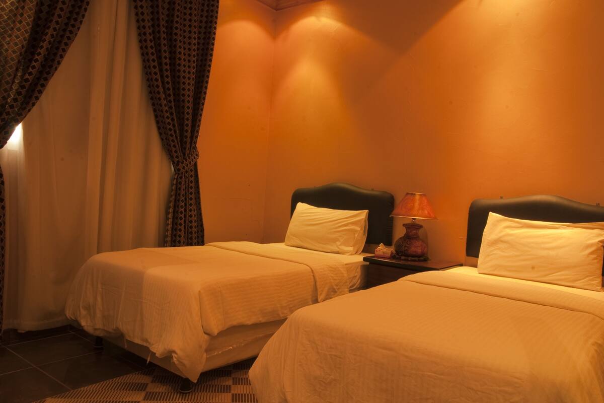 A hotel room with two single beds, a nightstand with a lamp, and a window with curtains.