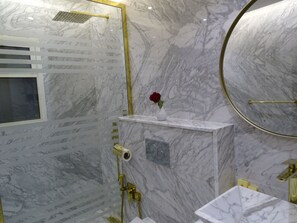 Ein Badezimmer mit Marmorwänden, einem goldfarbenen Duschkopf, einem weißen Waschbecken und einer Vase mit einer roten Rose.