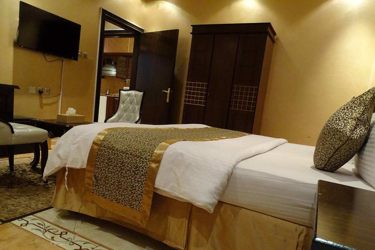 A hotel room with a bed, a television mounted on the wall, a wardrobe, and a desk with a chair.