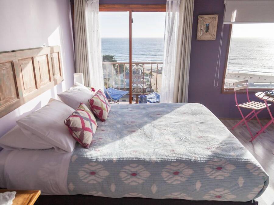 Un dormitorio con cama, almohadas, vista al mar, un balcón y una mesa con sillas.