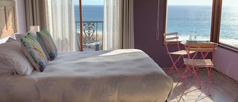 A bedroom with a bed, a window with a view of the sea, a small table with chairs, and a wooden floor.