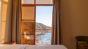 Quarto de hotel com cama, uma cadeira e vista para uma paisagem montanhosa através de uma janela com cortinas.