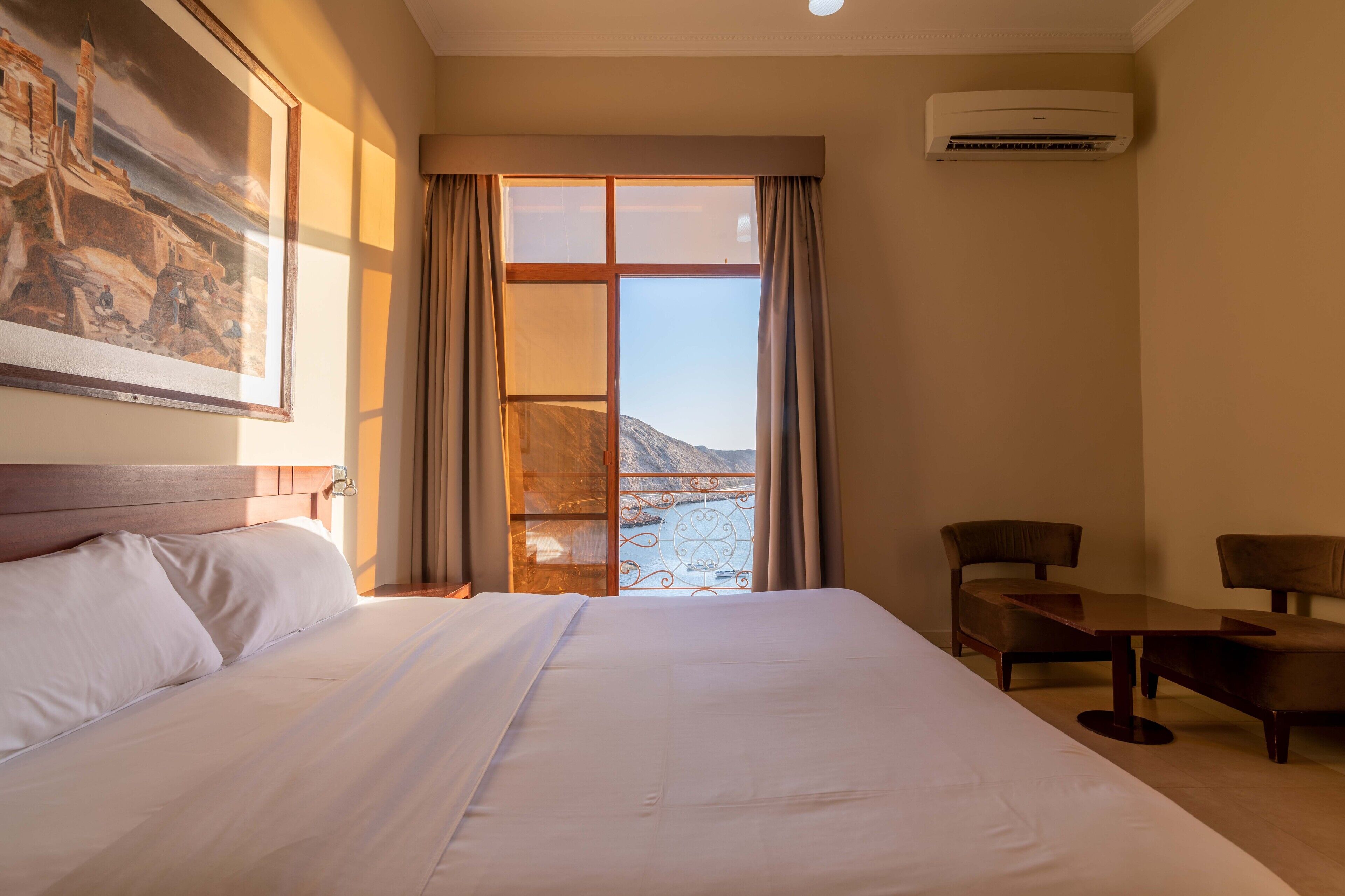 Quarto de hotel com uma cama grande, uma escrivaninha, duas cadeiras, uma janela com cortinas e um ar condicionado.