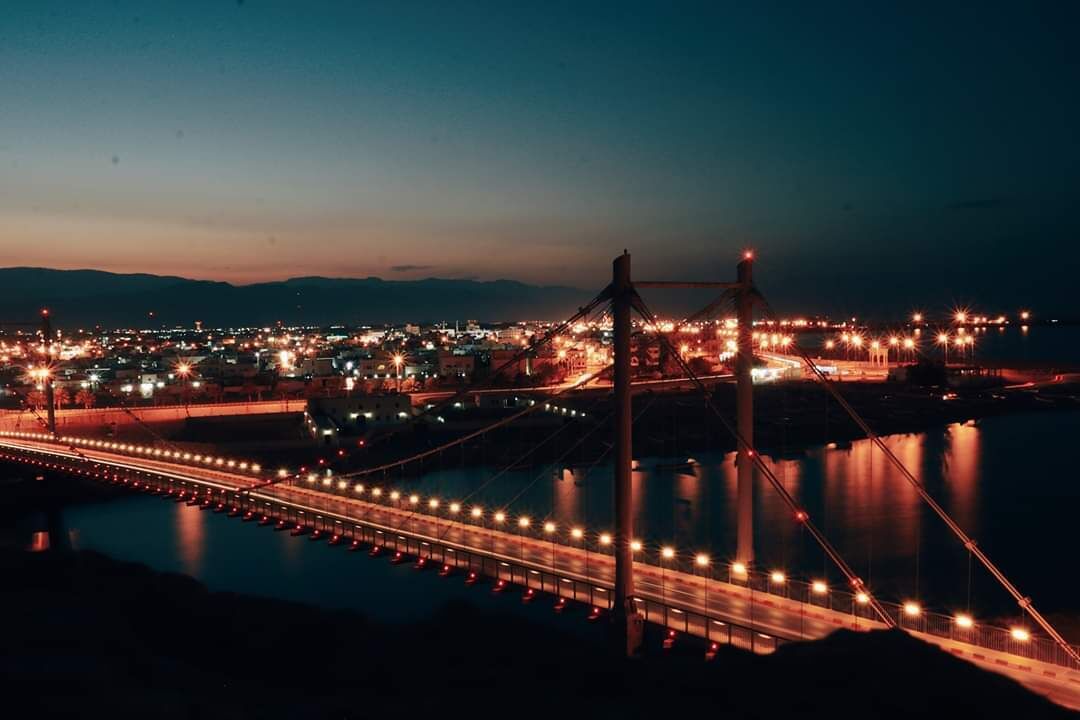 A nighttime view of a bridge with lights reflecting on the water, surrounded by a cityscape.