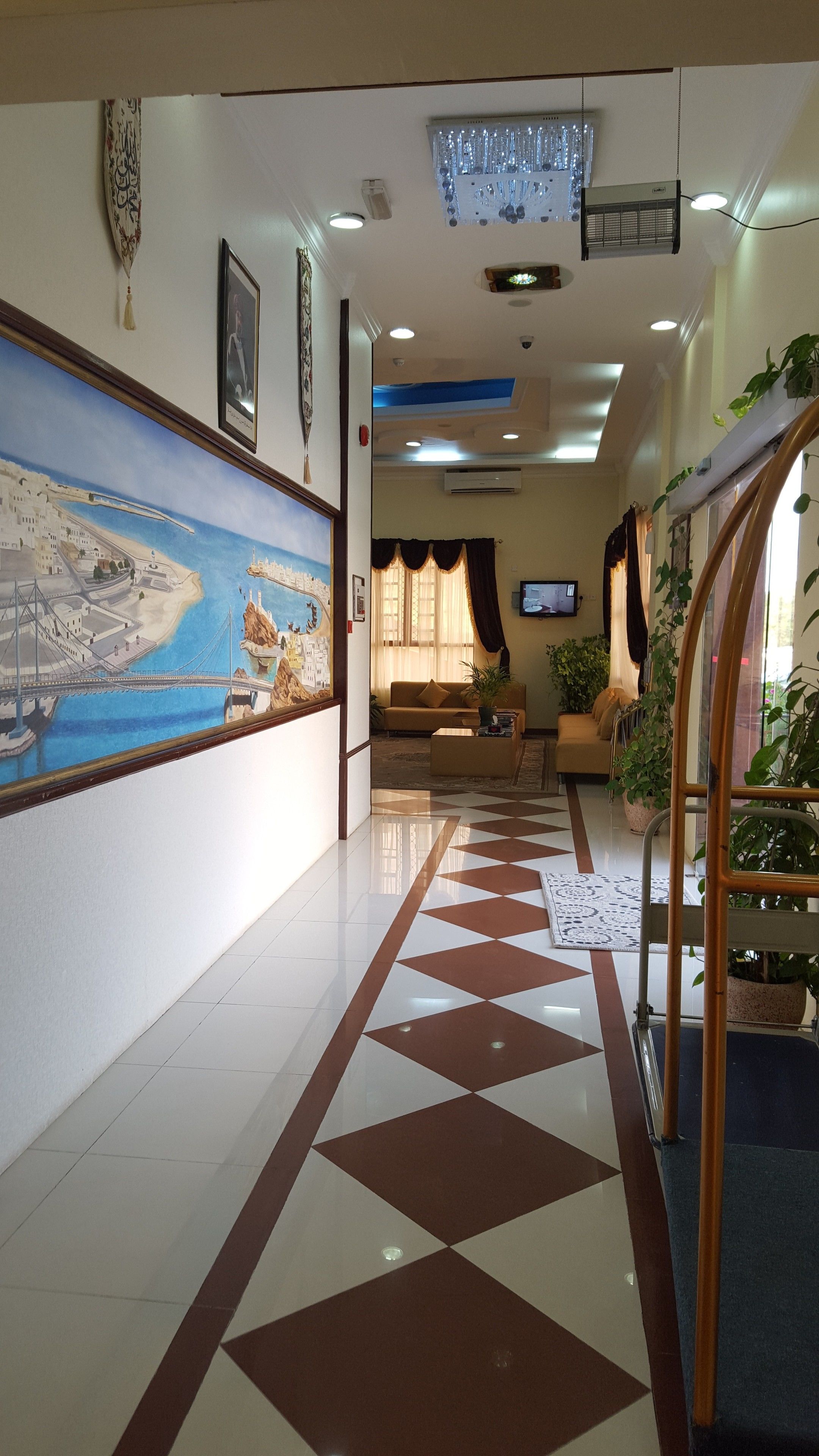 A hotel lobby with a tiled floor, a large painting of a coastal scene, a sofa, and a television.