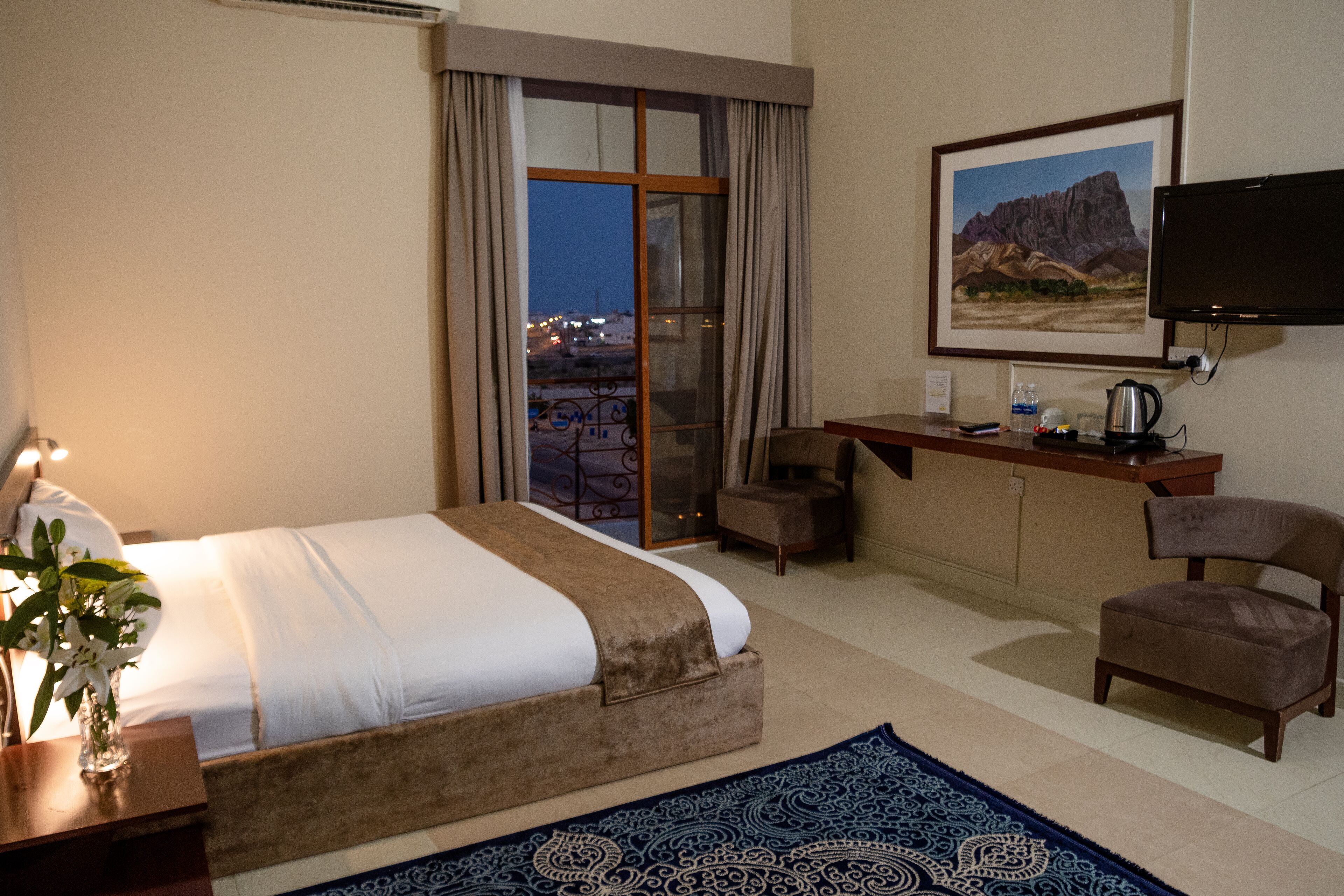A hotel room with a bed, a desk, a chair, a TV, and a view of the city at dusk.