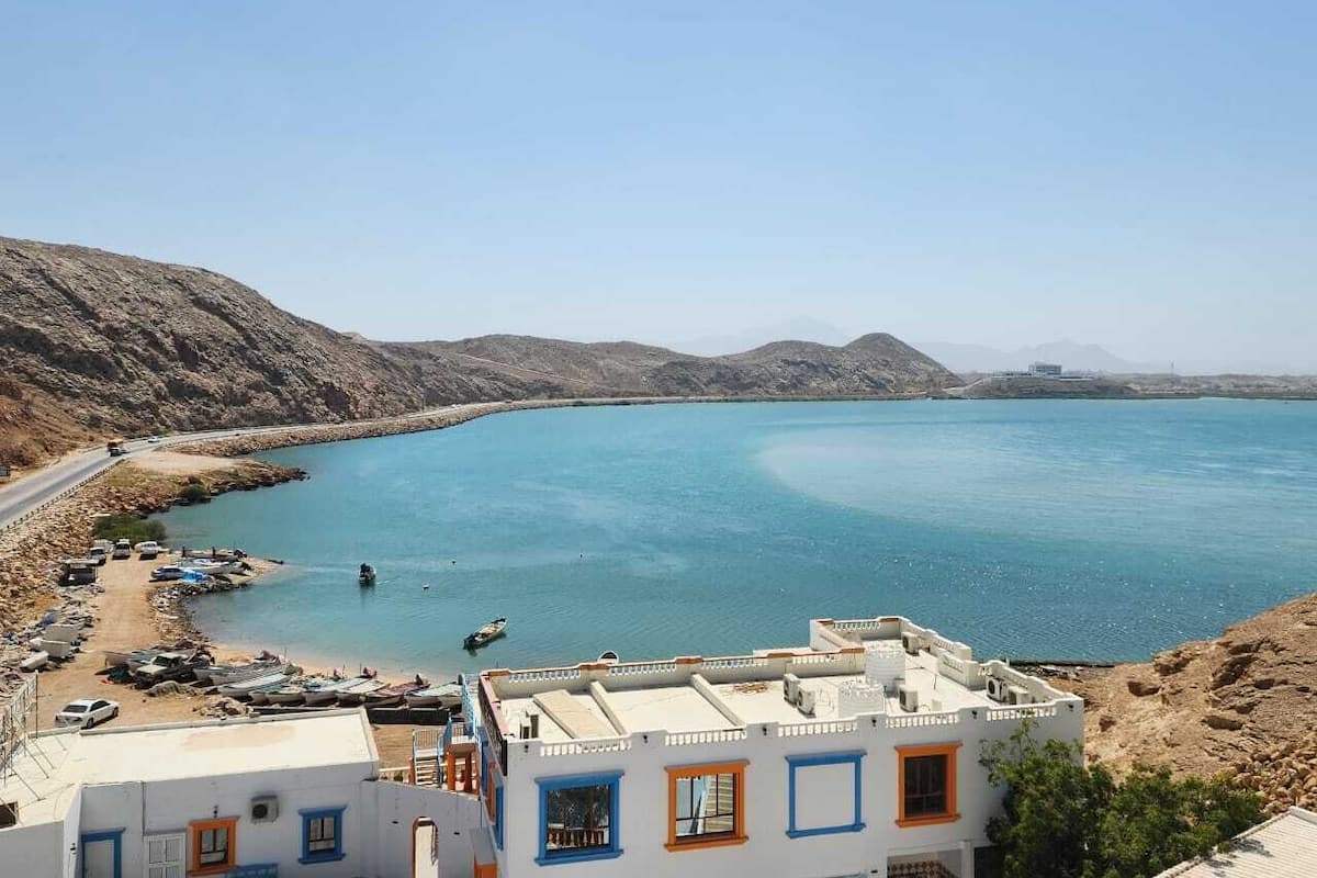 A coastal town with a clear blue sea, buildings with blue and orange accents, and a road running alongside the water.