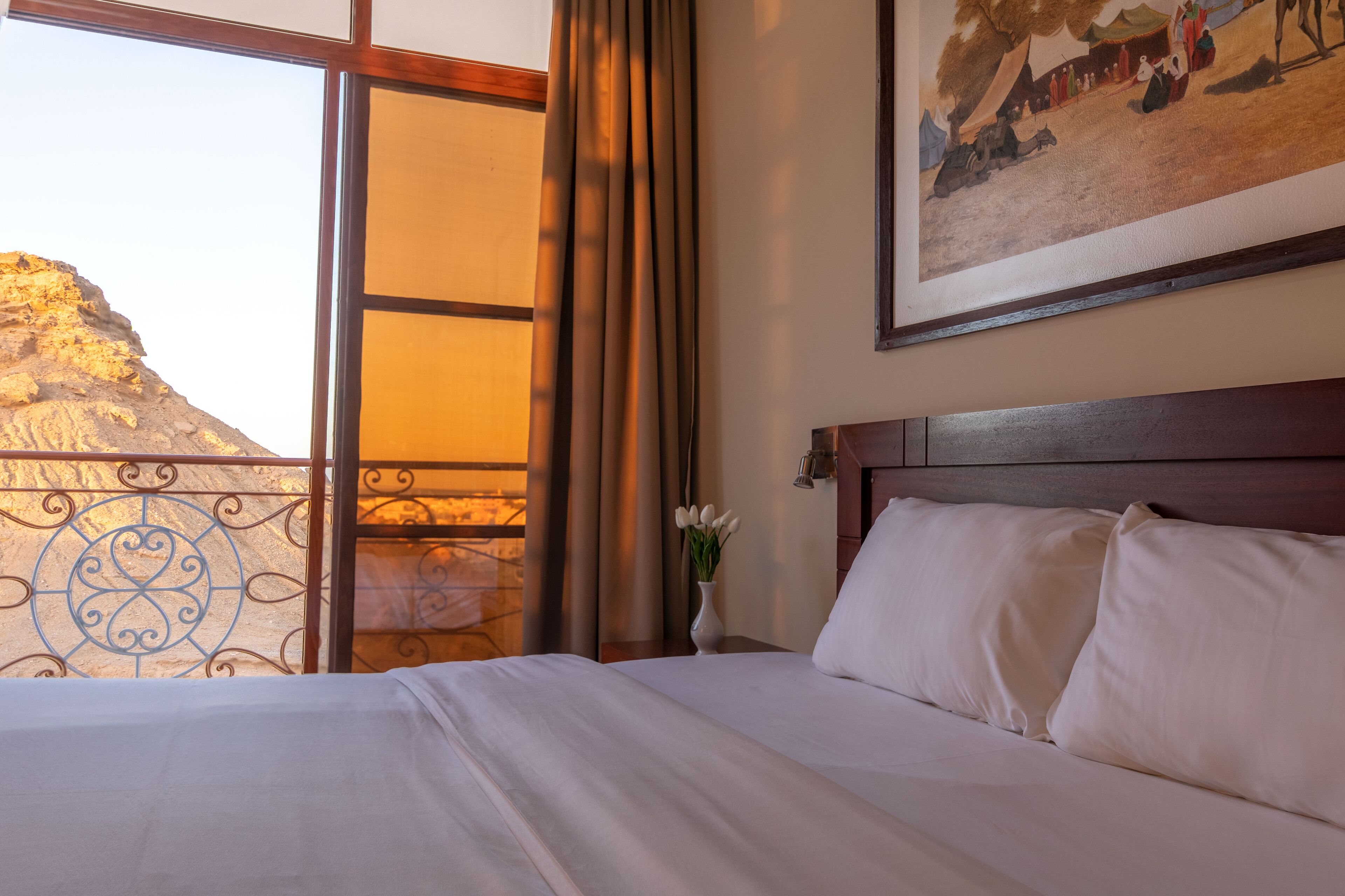 A hotel room with a bed, a painting on the wall, and a view of a mountain through a window.