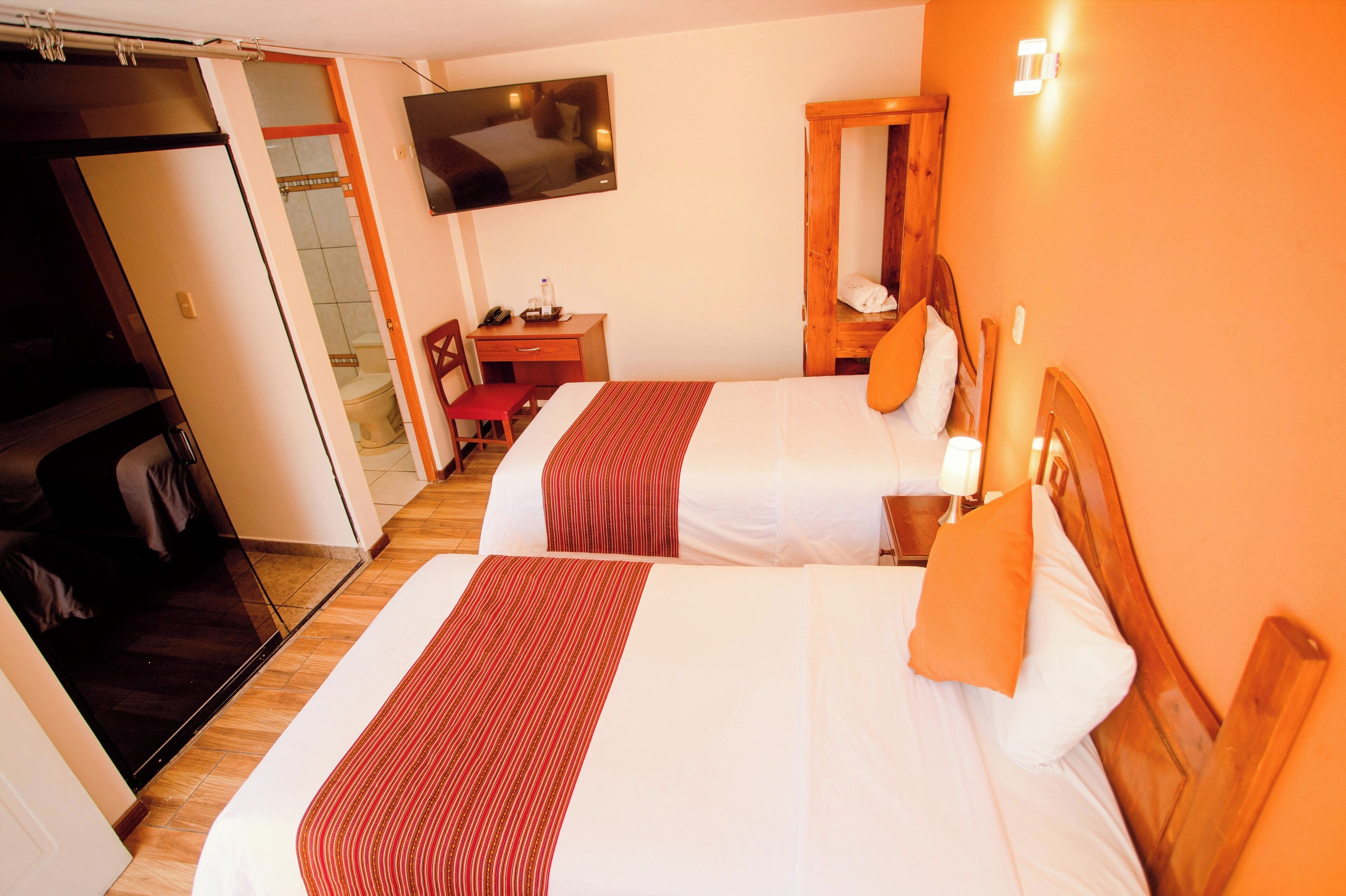 Habitación de hotel con dos camas, un televisor montado en la pared, un armario de madera y un escritorio con una silla.