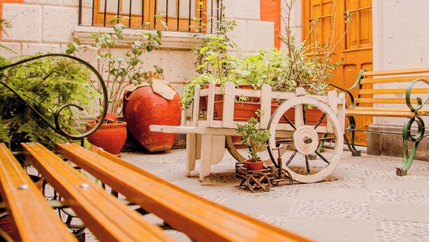 A courtyard with wooden benches, a laundry service sign, a wooden cart with potted plants, and a large orange door. - Hostal Villa Sillar (Arequipa)