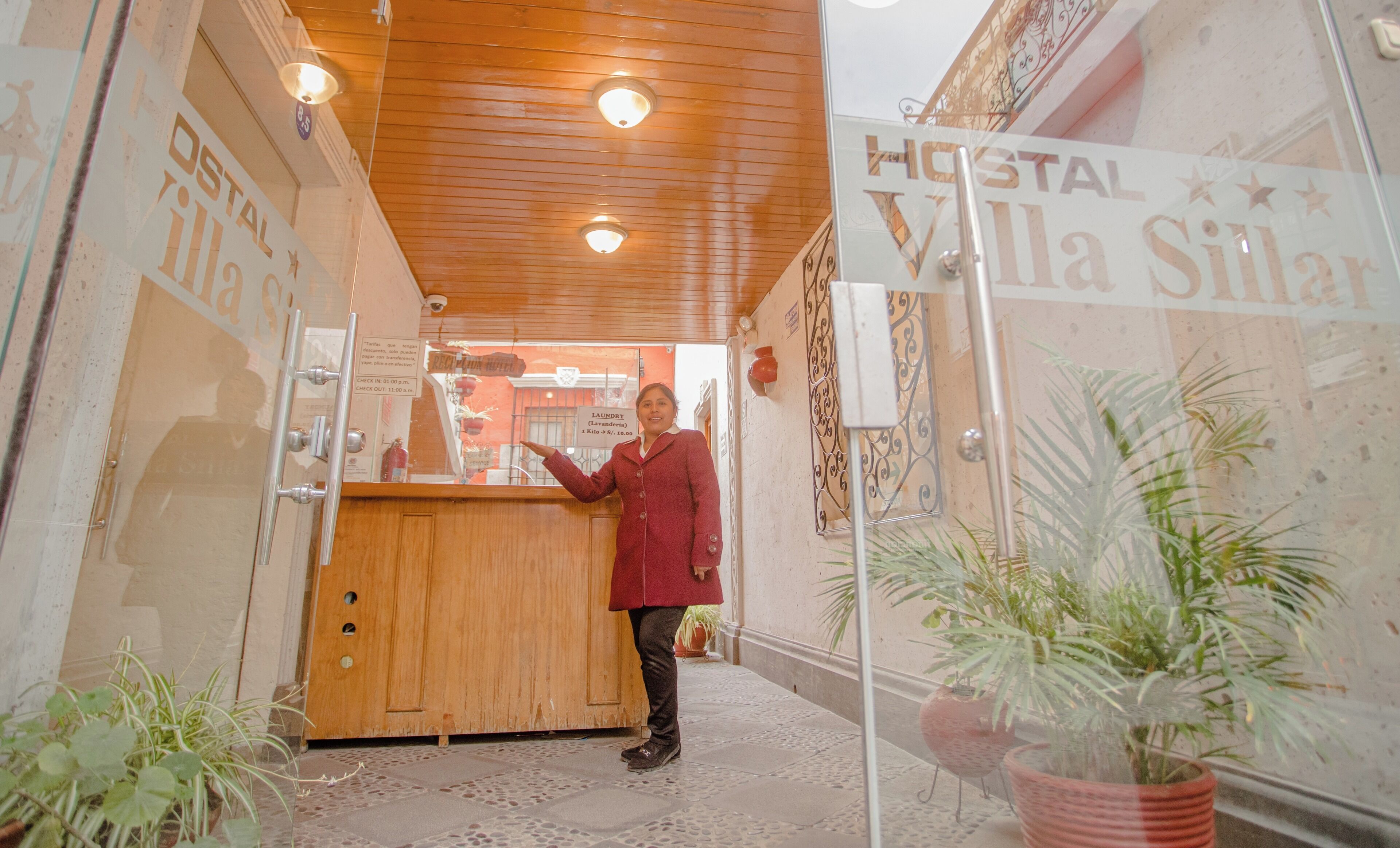 A hotel reception area with a wooden reception desk, a sign reading "HOSTAL Villa Sillar", and potted plants.