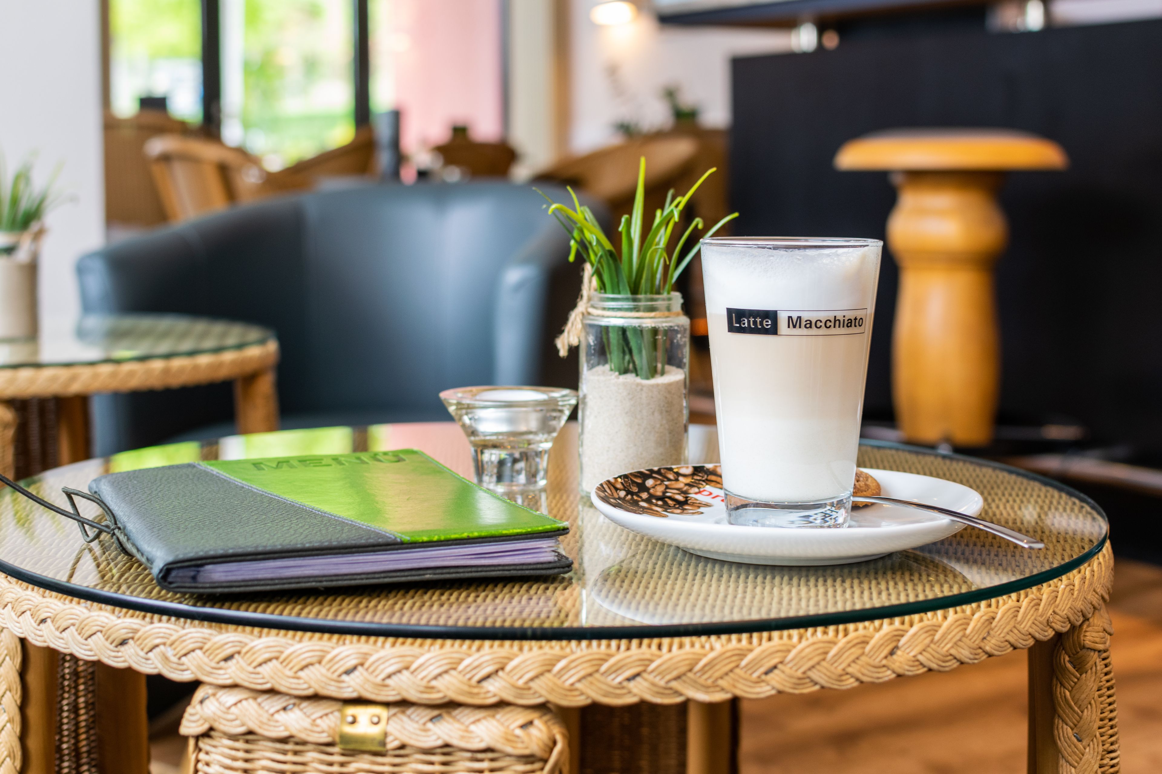 Taula amb un got de latte macchiato, un quadern, un got petit i un botic decoratiu amb plantes verdes.