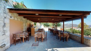 An outdoor patio with wooden tables and chairs, stone walls, and a view of the countryside. - Nana Apartments (Faistos)