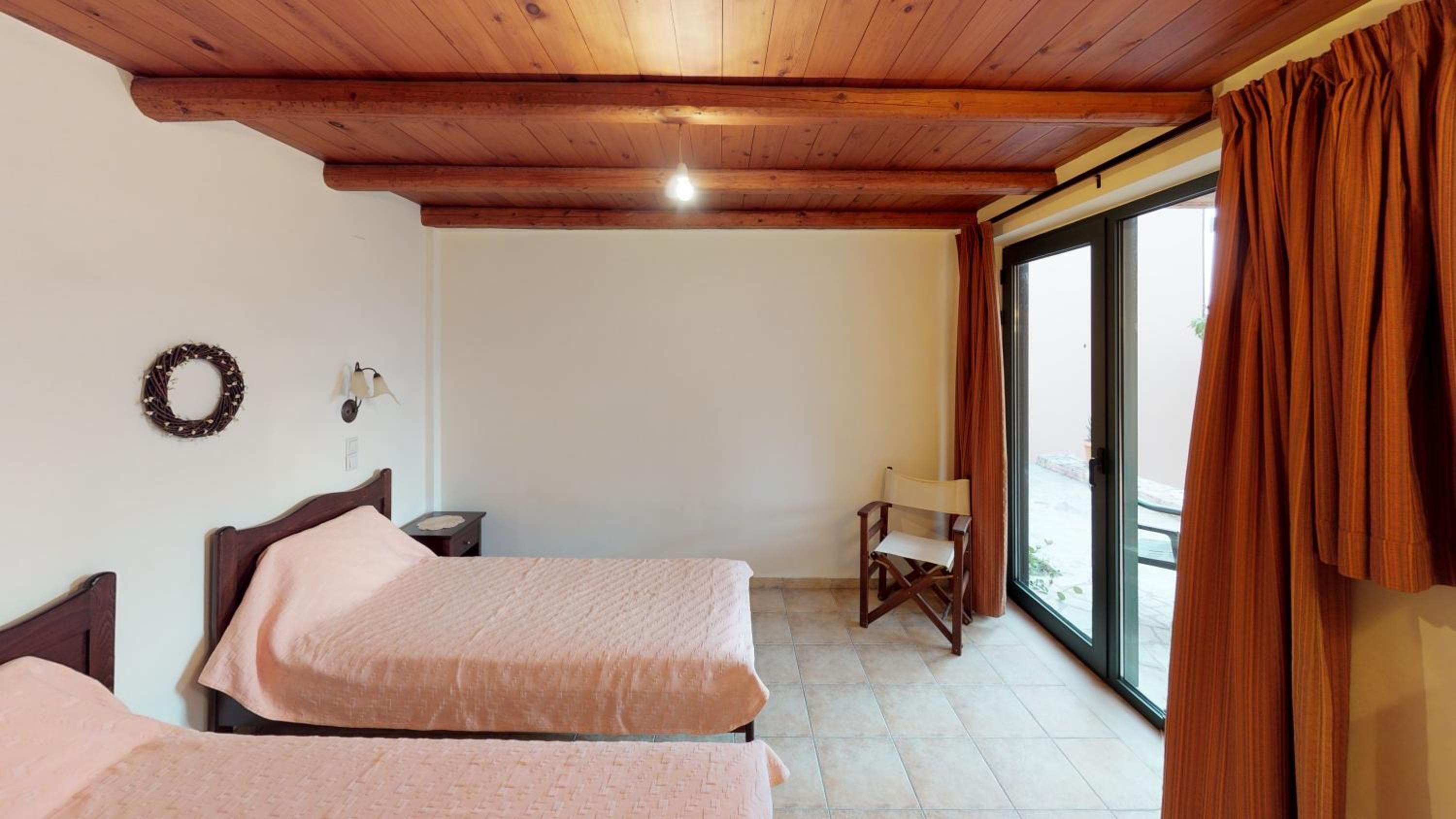 A bedroom with two beds, a small table, a chair, a wall-mounted lamp, and a window with curtains.