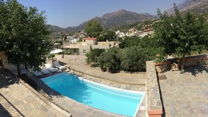 A swimming pool with lounge chairs, surrounded by stone walls and greenery, with a view of a hillside town in the background. - Nana Apartments (Faistos)