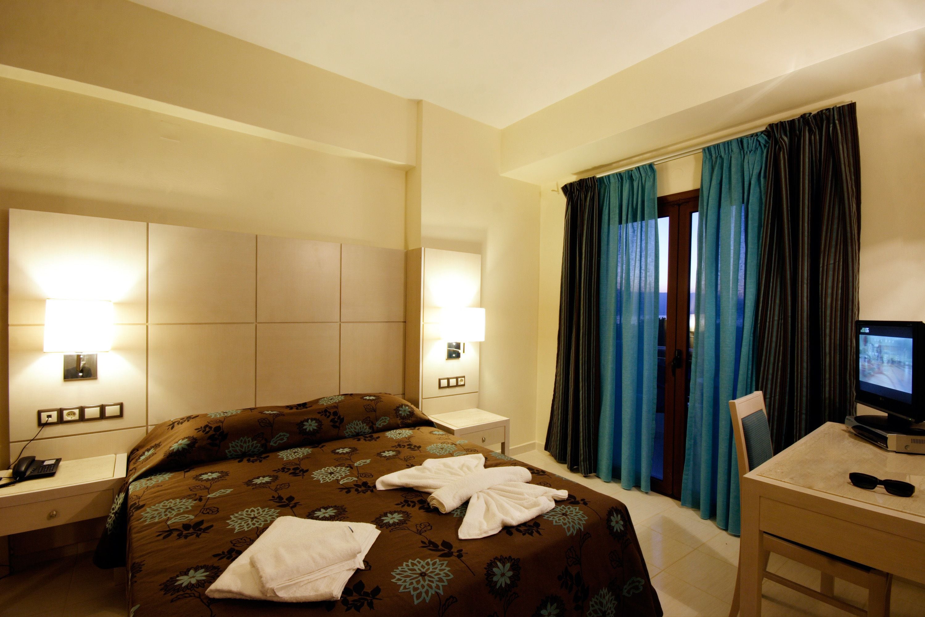 Una habitación de hotel con cama, lámparas de noche, escritorio y televisor, y cortinas.