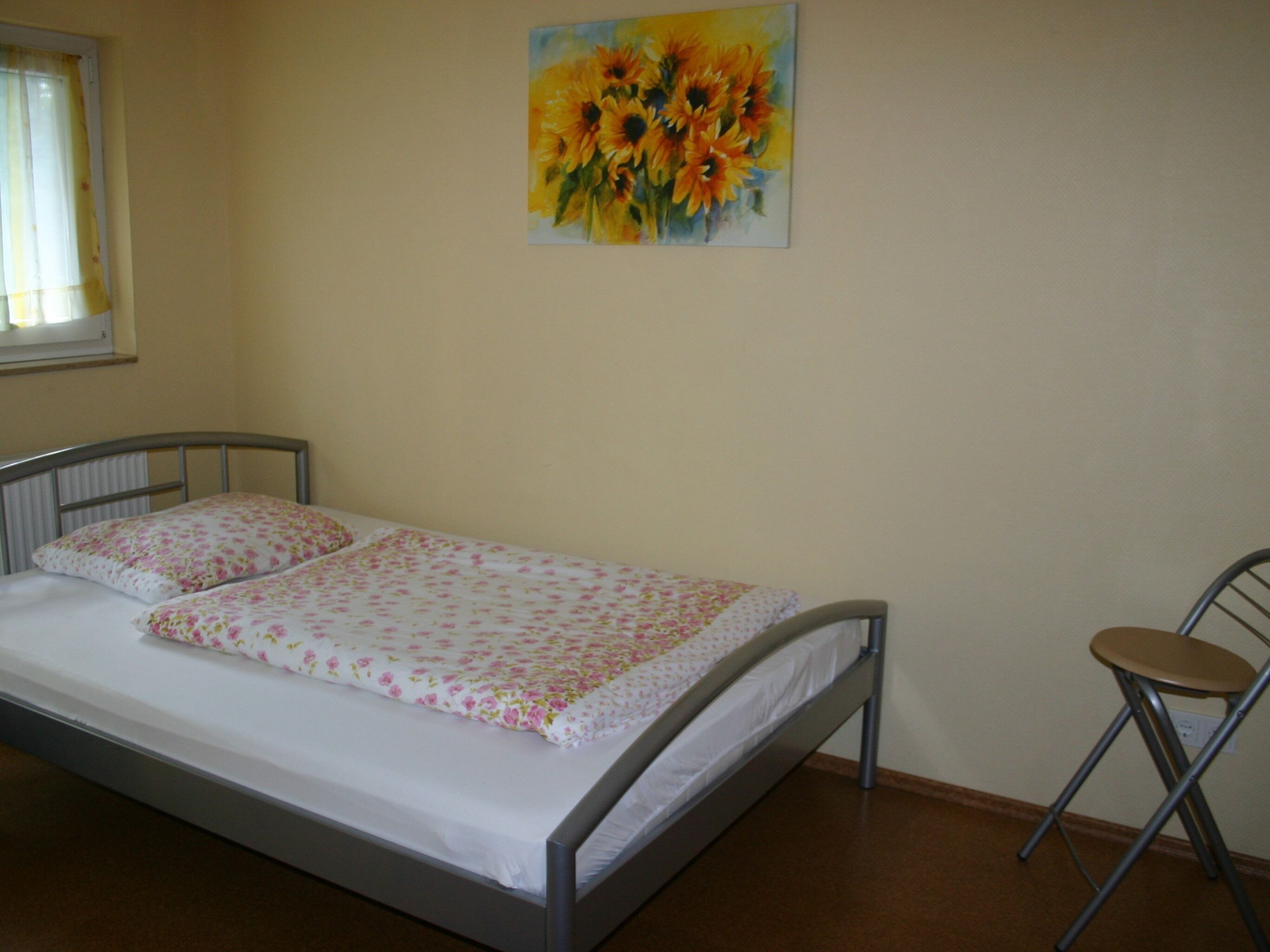 A single bed with a floral bedspread and two pillows, a small stool, and a painting of sunflowers on the wall.