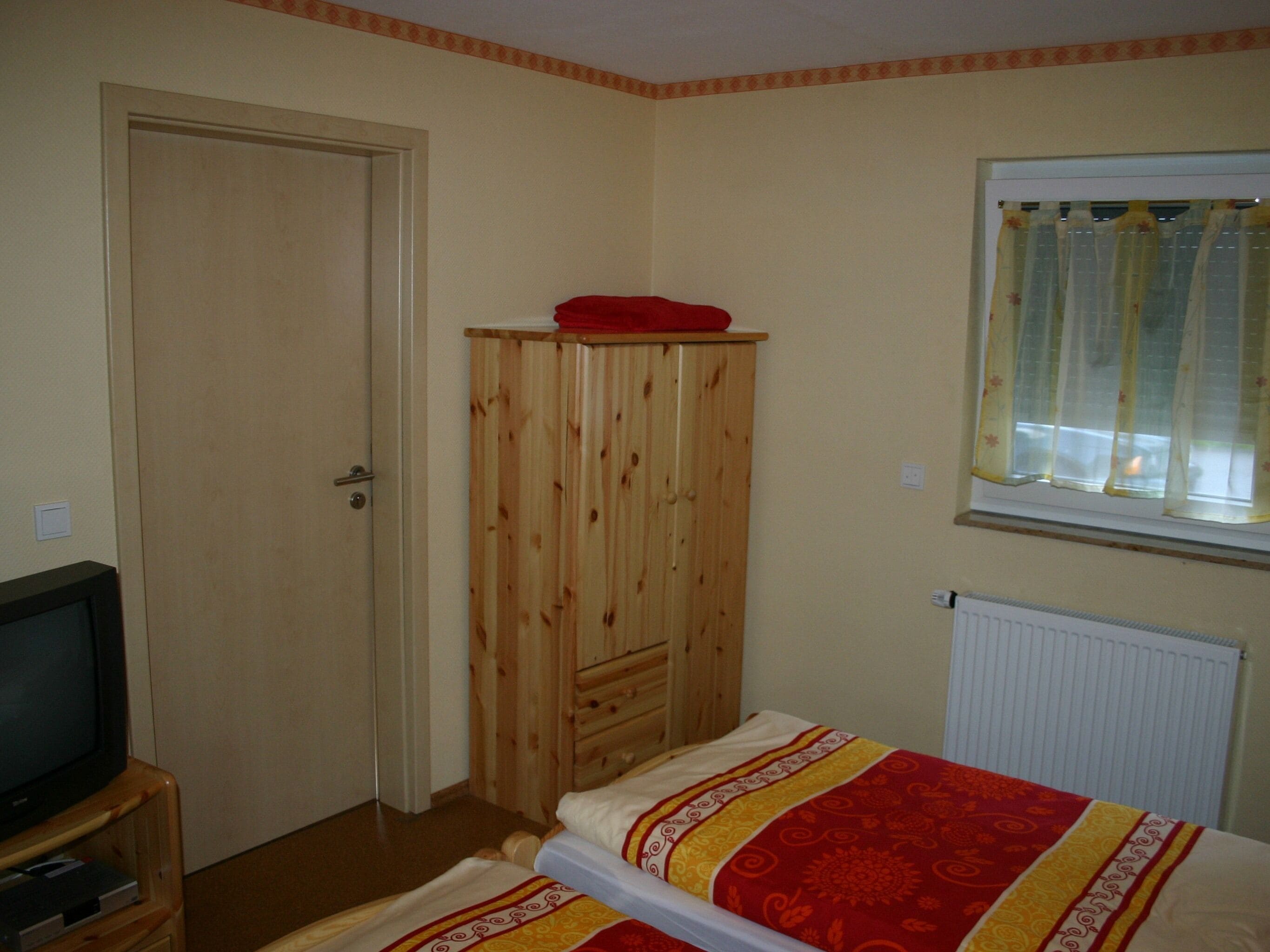 Een slaapkamer met een houten garderobe, een bed met een bedovertrek met patroon, een televisie op een houten dressoir en een raam met gordijnen.