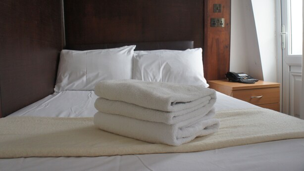 A neatly made bed with white linens and a folded white towel on top, a wooden nightstand with a lamp and a phone, and a window with a view of the outside. - Plaza London Hotel (London)