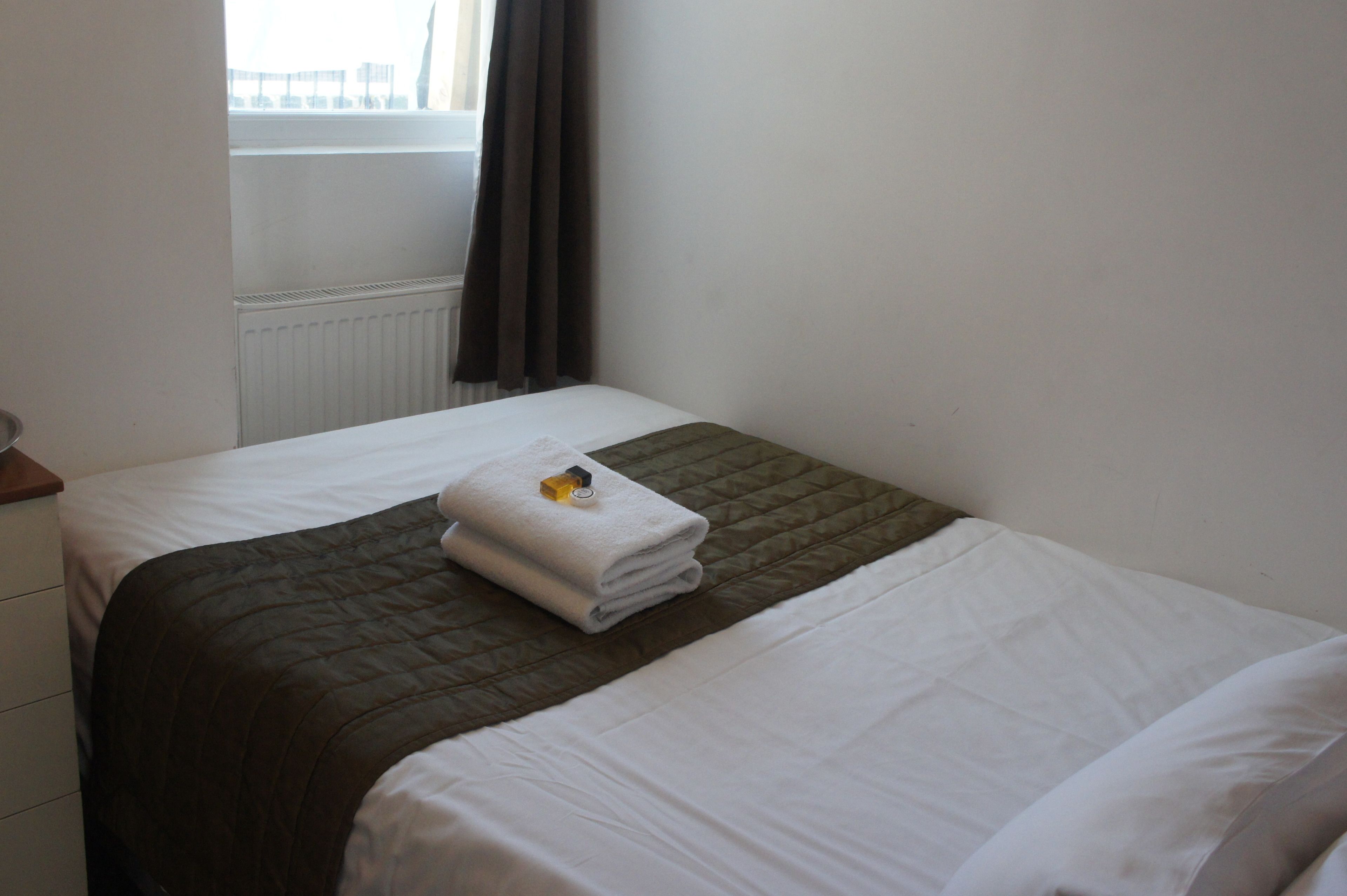 Una cama bien hecha con una toalla blanca doblada y un objeto amarillo pequeño encima, una ventana con cortinas oscuras y un radiador blanco.