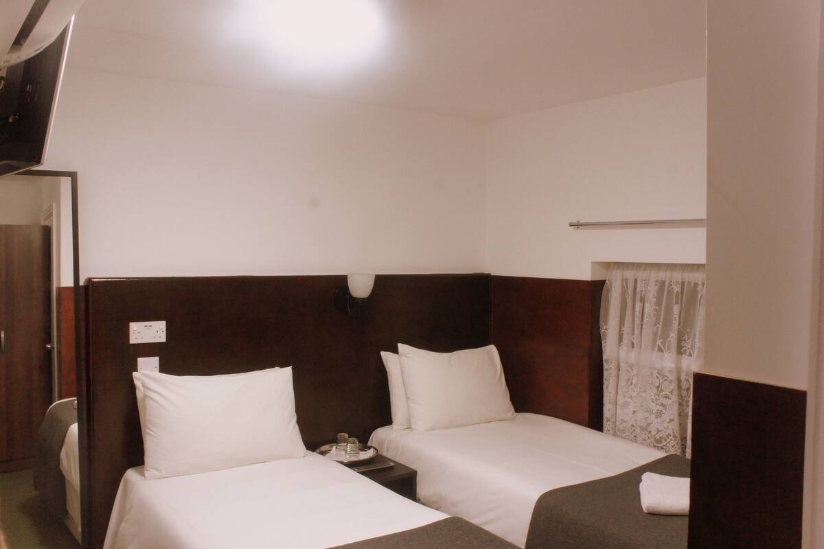 A hotel room with two single beds, a small table with a tray of items, a mirror, and a window with sheer curtains.