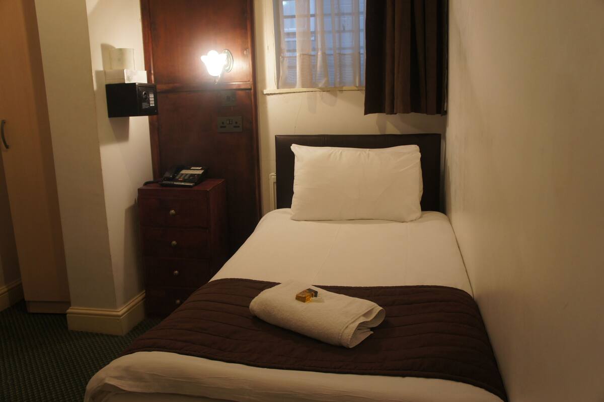 A hotel room with a bed, a bedside table with a telephone, a nightstand, a window with curtains, and a wall-mounted light.