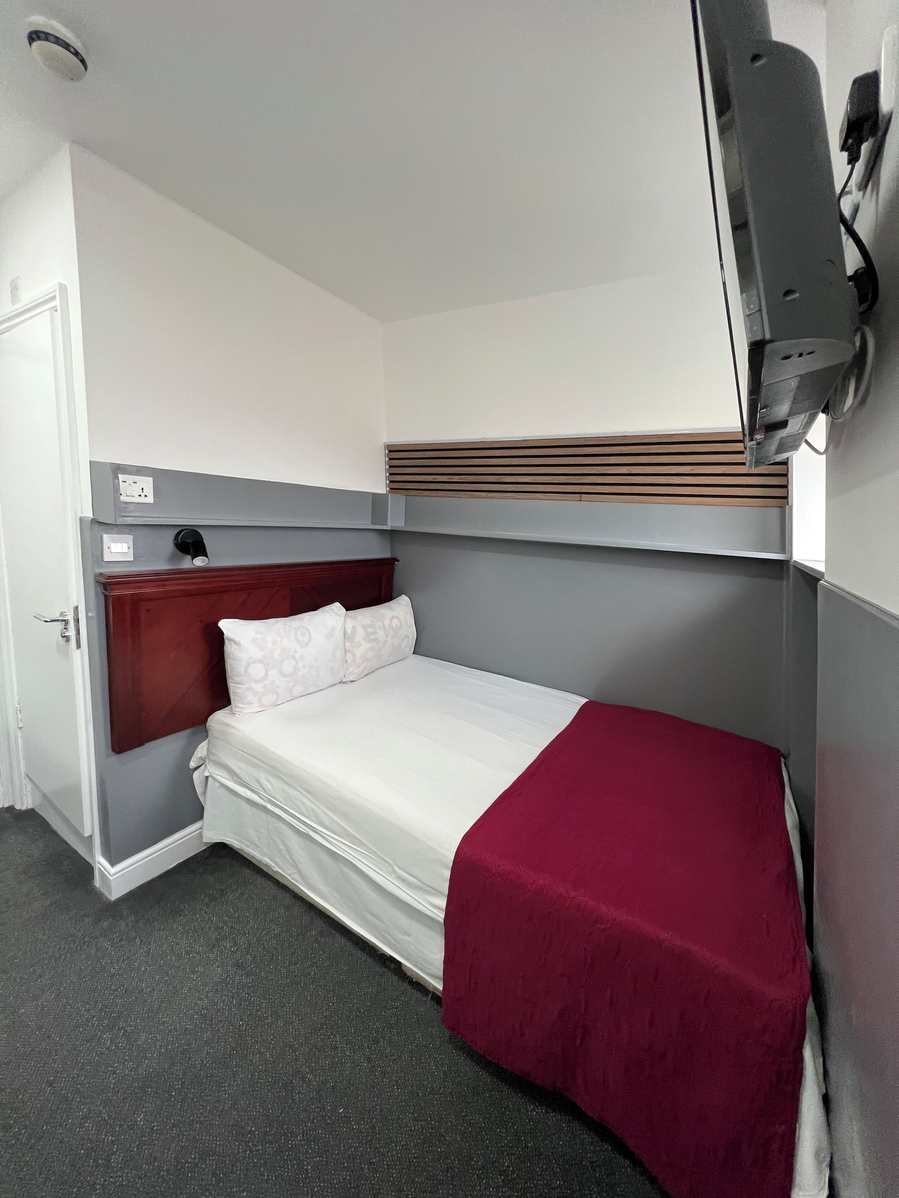 Une chambre d’hôtel de taille modeste, comprenant un lit simple, une tête de lit en bois, un plaid rouge et un petit bureau avec une chaise.