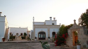 Une cour intérieure avec un bâtiment doté d’une porte centrale et de fenêtres en arc de cercle, entouré de plantes en pot et d’un cactus.