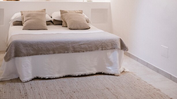 Une chambre à coucher avec un plafond voûté, une lucarne, un lit recouvert d’une couvre-lit blanche et grise, deux lampes de chevet et un vase contenant des plantes séchées sur la table de chevet.