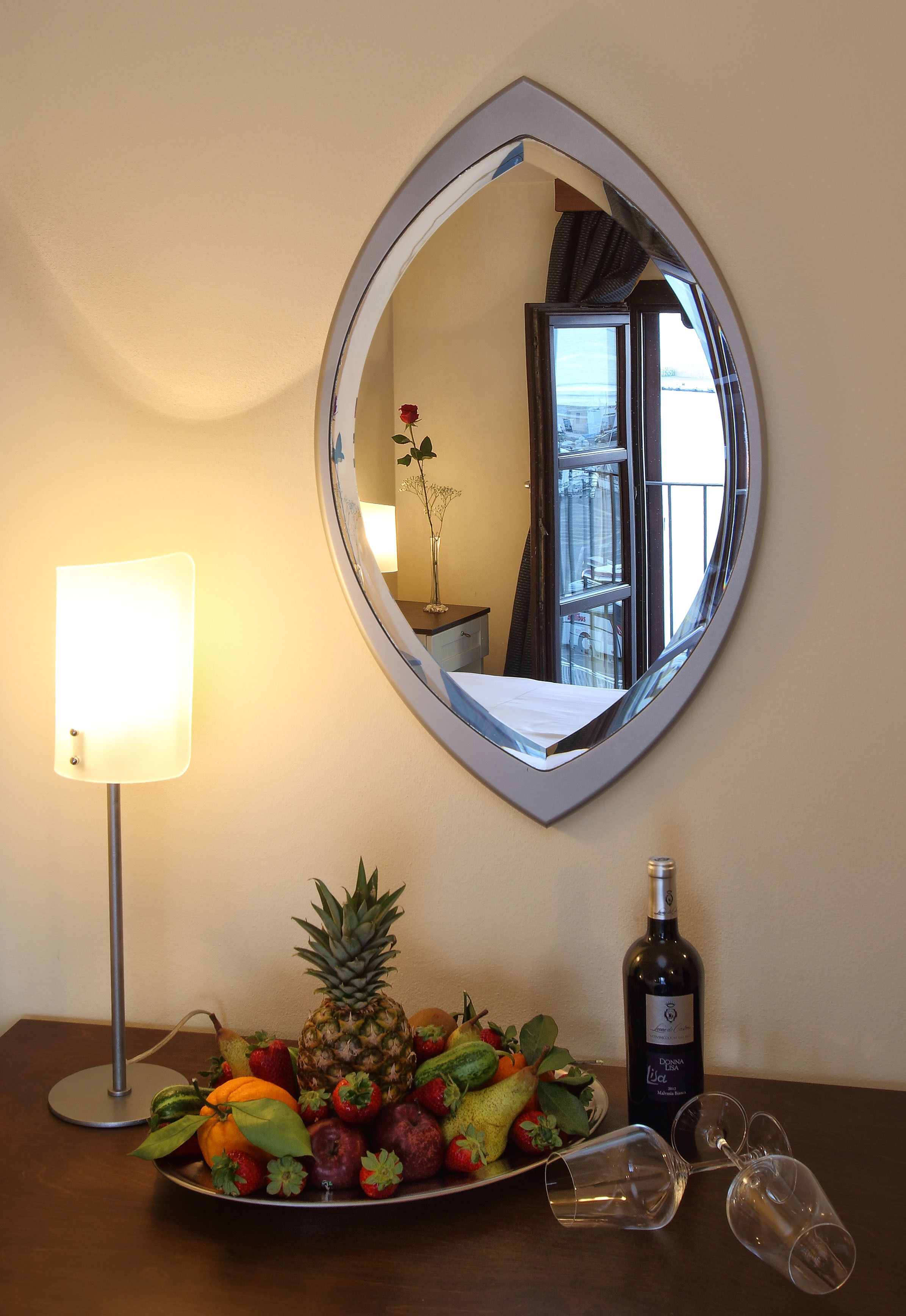 Een hotelkamer met een ronde spiegel, een fruitplateau, een fles wijn en een bedlamp.