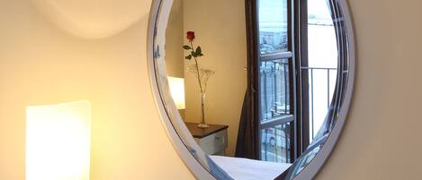 A hotel room with a round mirror, a fruit platter, a bottle of wine, and a bedside lamp.