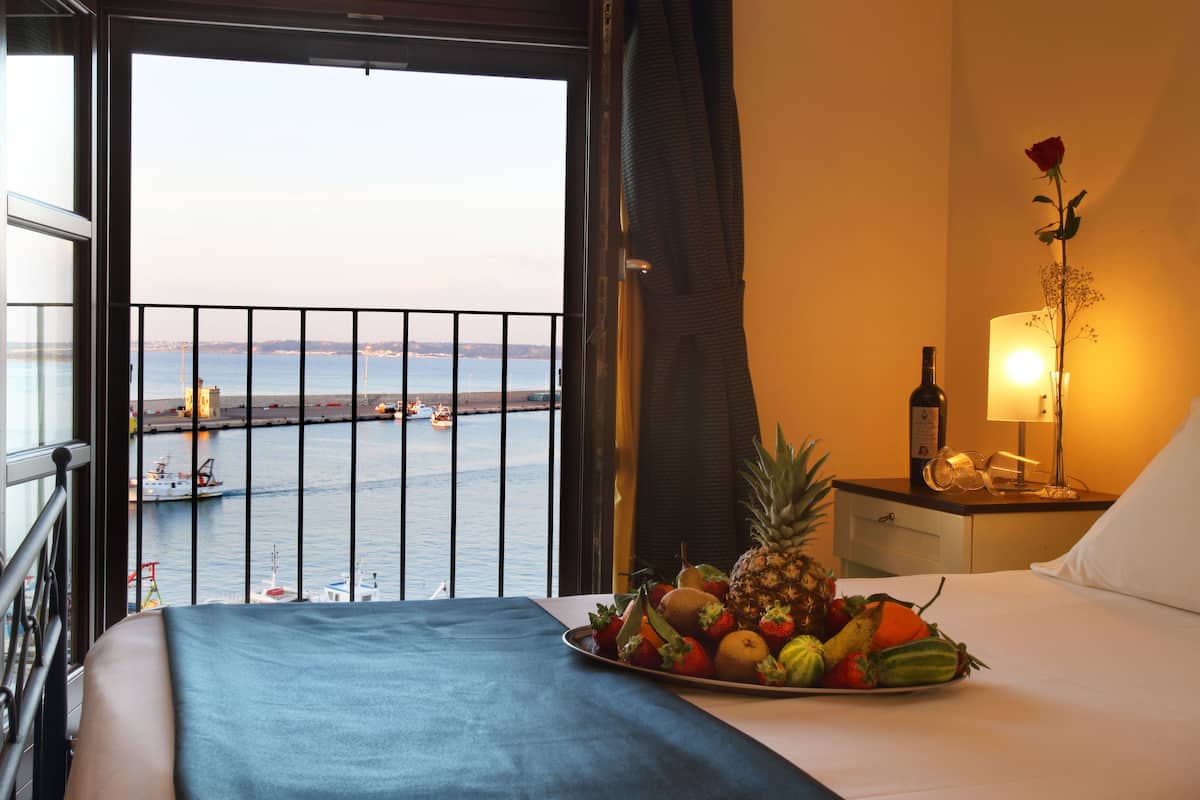 A hotel room with a balcony overlooking a body of water, a bed with a fruit basket, a nightstand with a lamp, and a rose decoration.