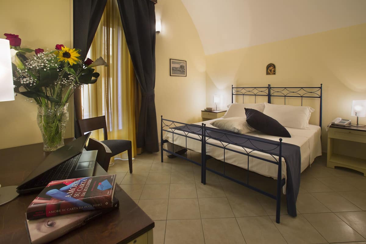 A bedroom with a bed, a desk with a laptop, a vase with flowers, and a bedside table with a lamp.