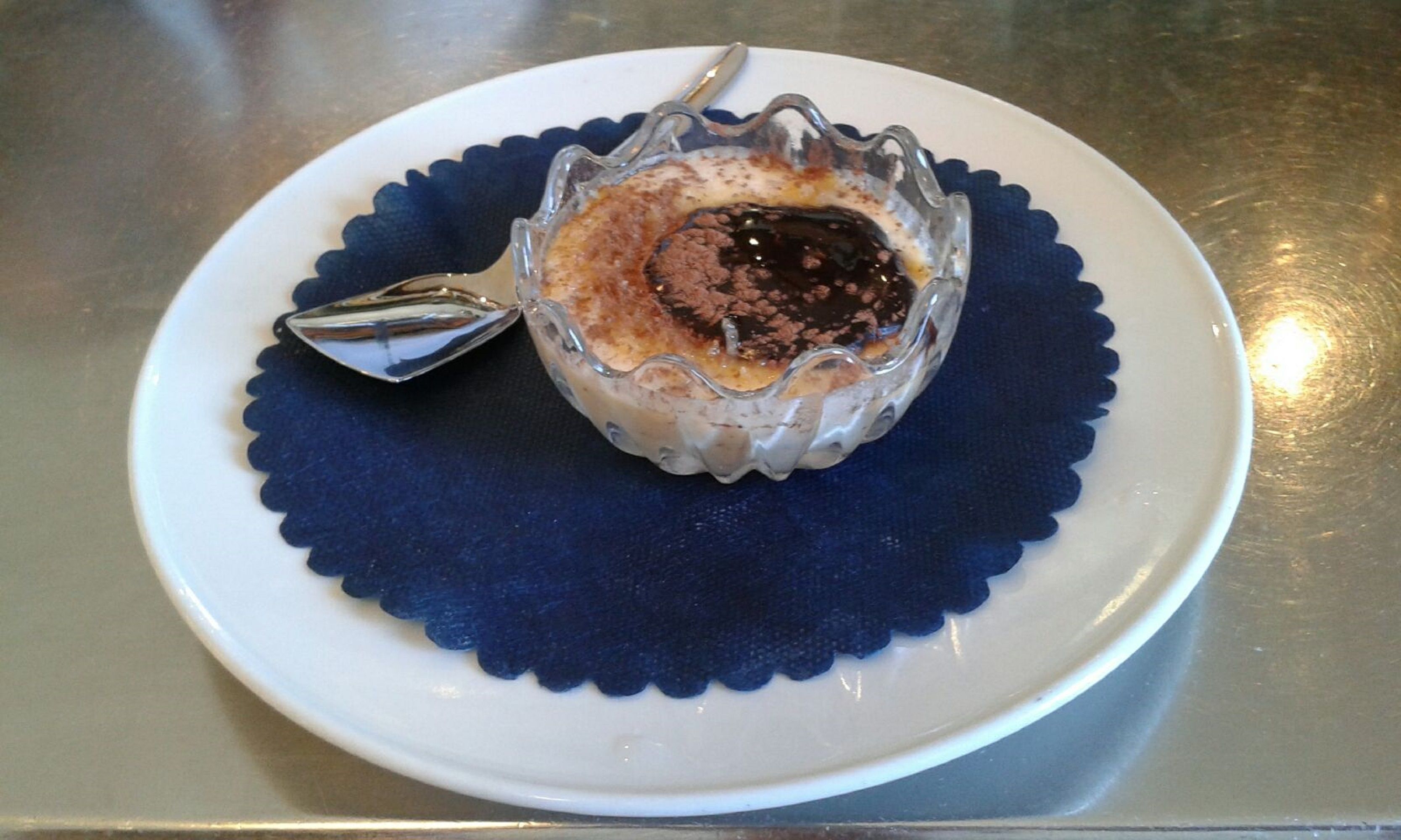 A dessert in a glass bowl on a white plate with a blue scalloped edge, placed on a metal surface.