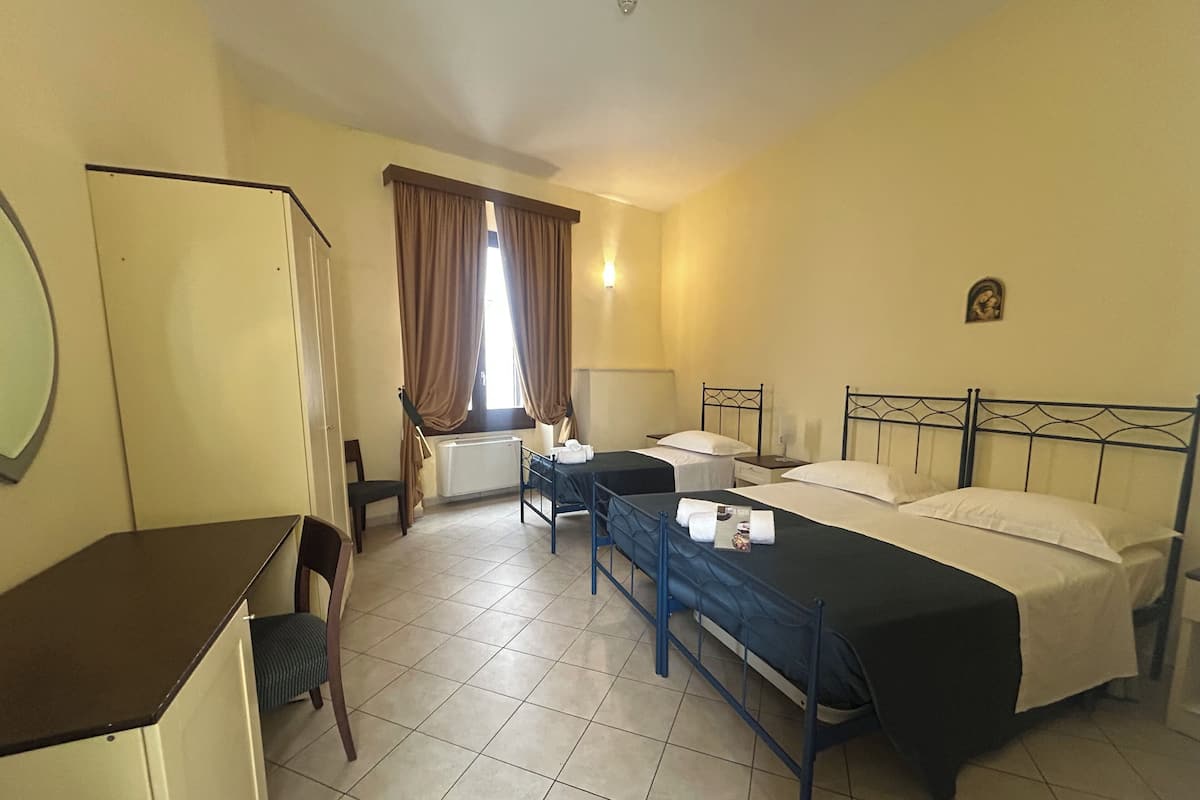 A hotel room with two beds, a desk, a chair, a radiator, and a window with curtains.