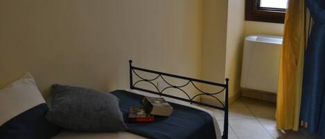 A single bed with a metal headboard, a bedside table with a book, and a window with curtains.