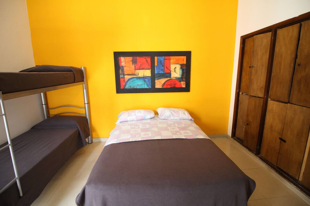 A room with a bunk bed, a single bed, a wooden wardrobe, and a colorful abstract painting on the wall.