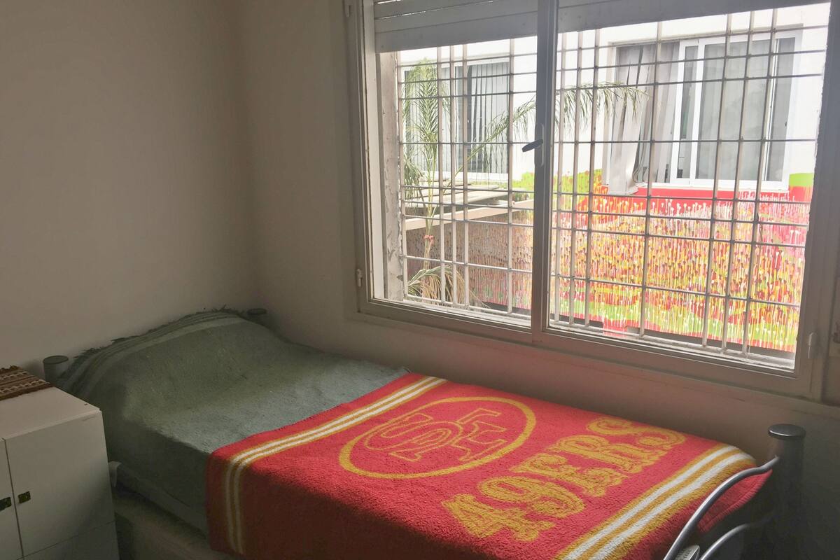 A small room with a single bed, a window with metal bars, and a view of a colorful outdoor area.