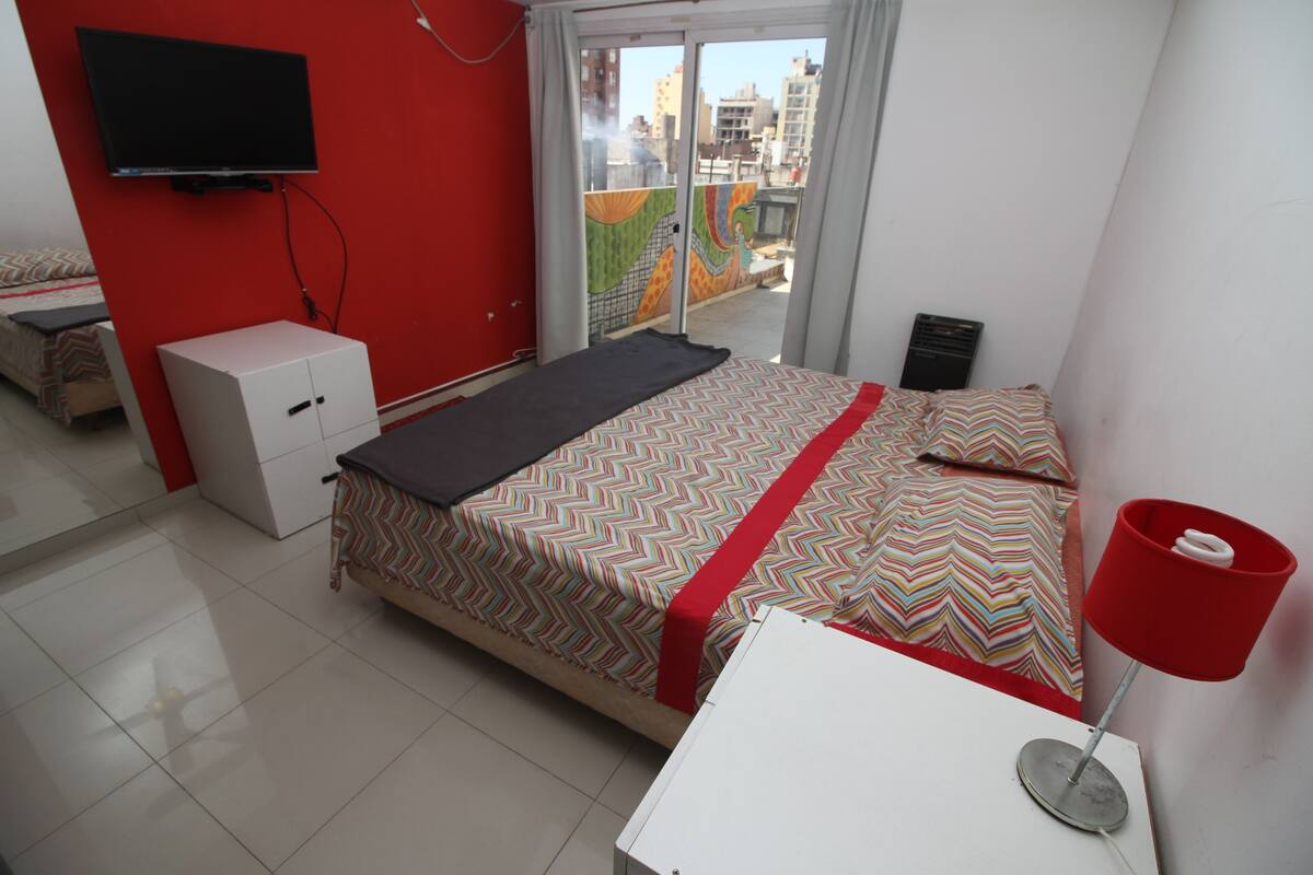 A bedroom with a bed, a desk, a TV, and a window with a view of buildings.