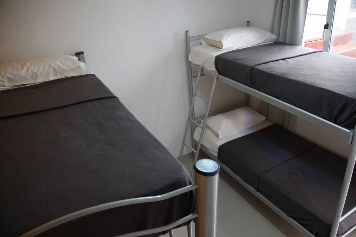 A dormitory room with bunk beds, a metal ladder, a trash can, and a window with curtains.