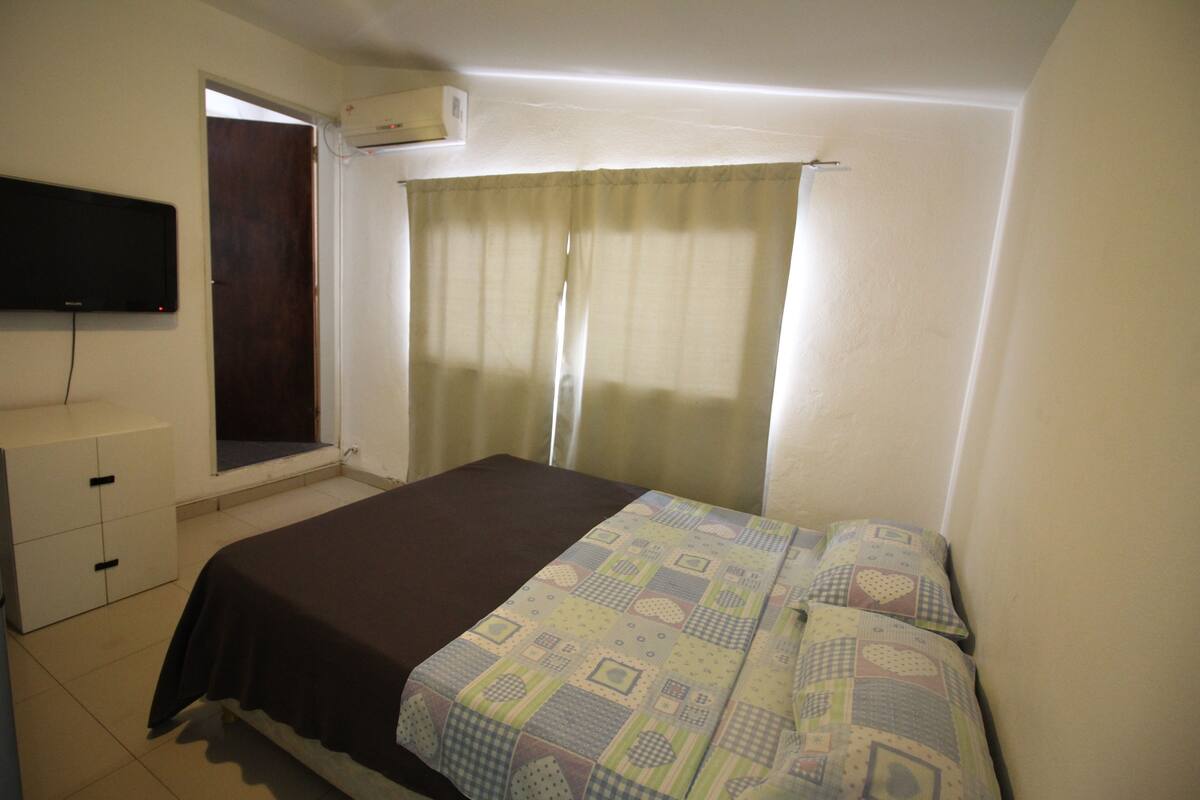 A bedroom with a bed, a window with curtains, a TV mounted on the wall, and a small dresser.