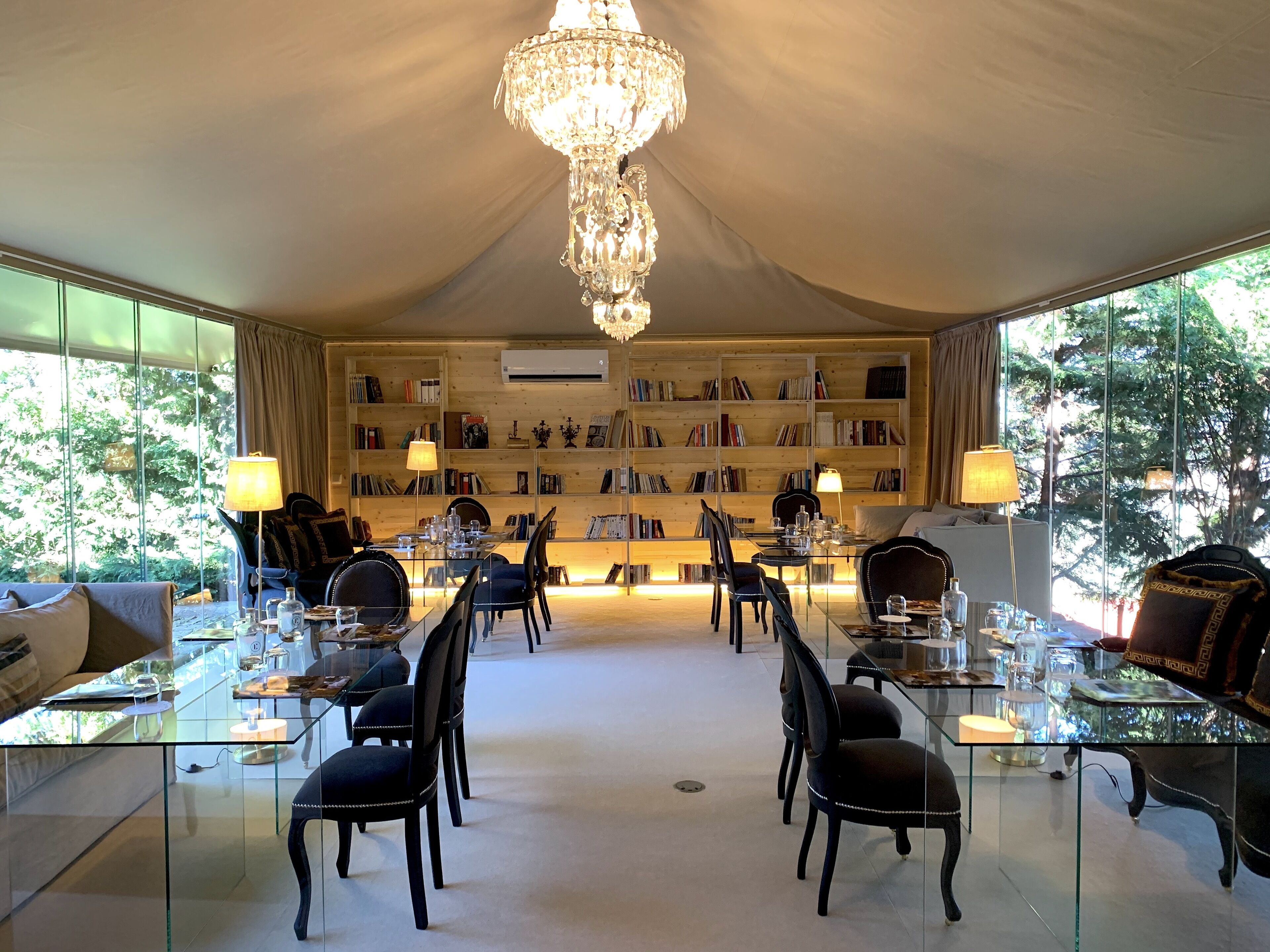 Una habitación lujosa con una gran araña, una mesa de comedor de cristal rodeada de sillas y una estantería llena de libros.