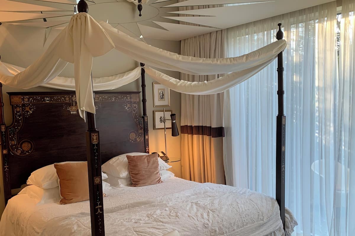 Una cama con dosel y pilares, cabecera de madera con intrincados grabados, mesita de noche con lámpara, y vista al exterior a través de cortinas translúcidas.