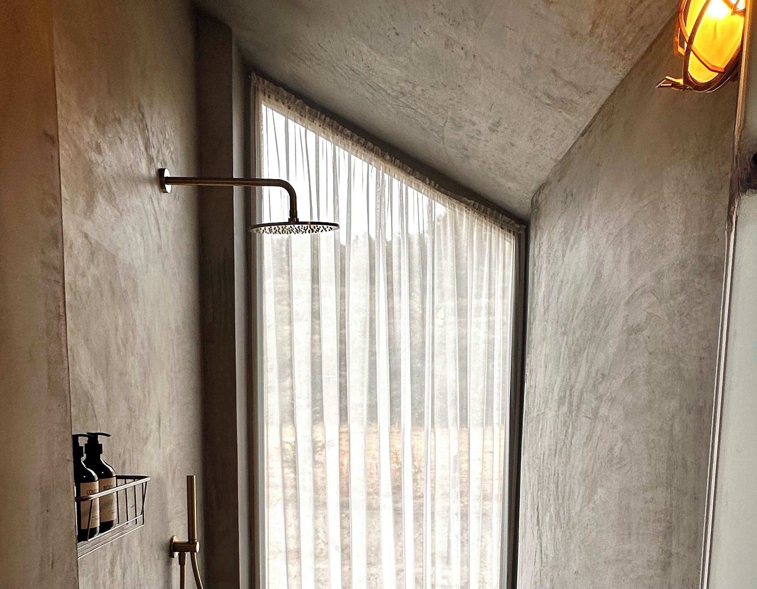 A modern bathroom with a shower, a window with sheer curtains, and a wall-mounted shelf with bottles.