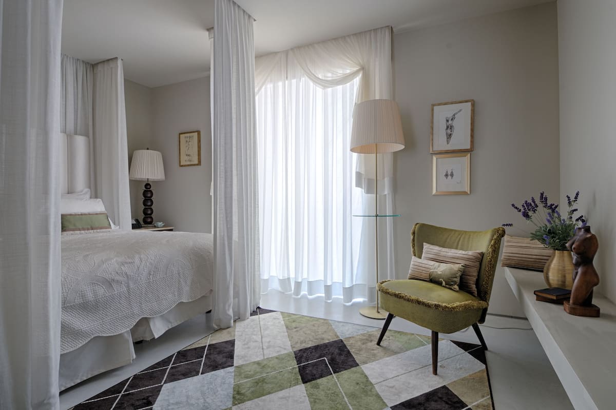 Un dormitorio con cama, una silla, una lámpara y ventana con cortinas translúcidas.