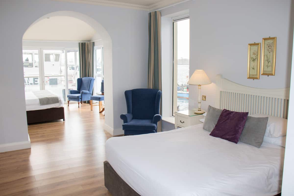 A hotel room with a large bed, a blue armchair, a small table, and a view of the city through a window.