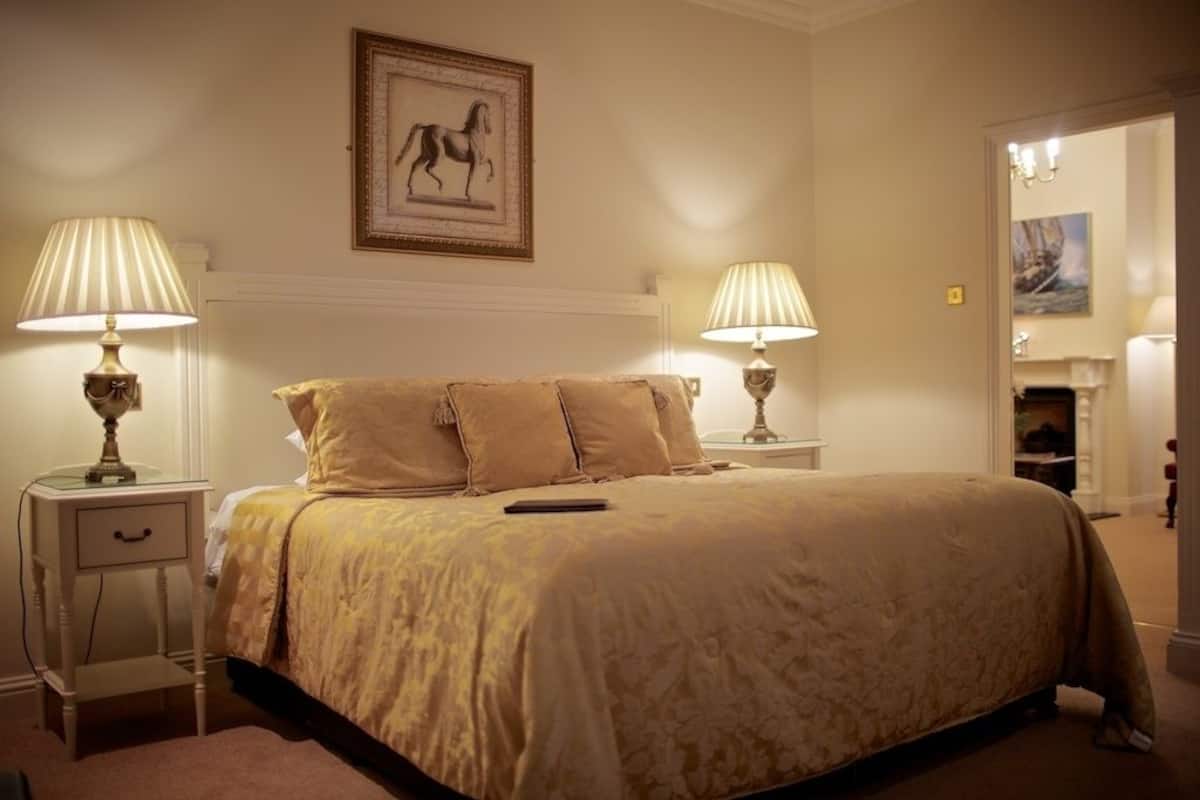 A bedroom with a large bed, two bedside tables with lamps, a framed picture on the wall, and a door leading to another room.