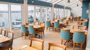 A restaurant with wooden tables and chairs, large windows overlooking a body of water, and a view of boats and buildings in the distance. - WatersEdge Hotel (Cobh)